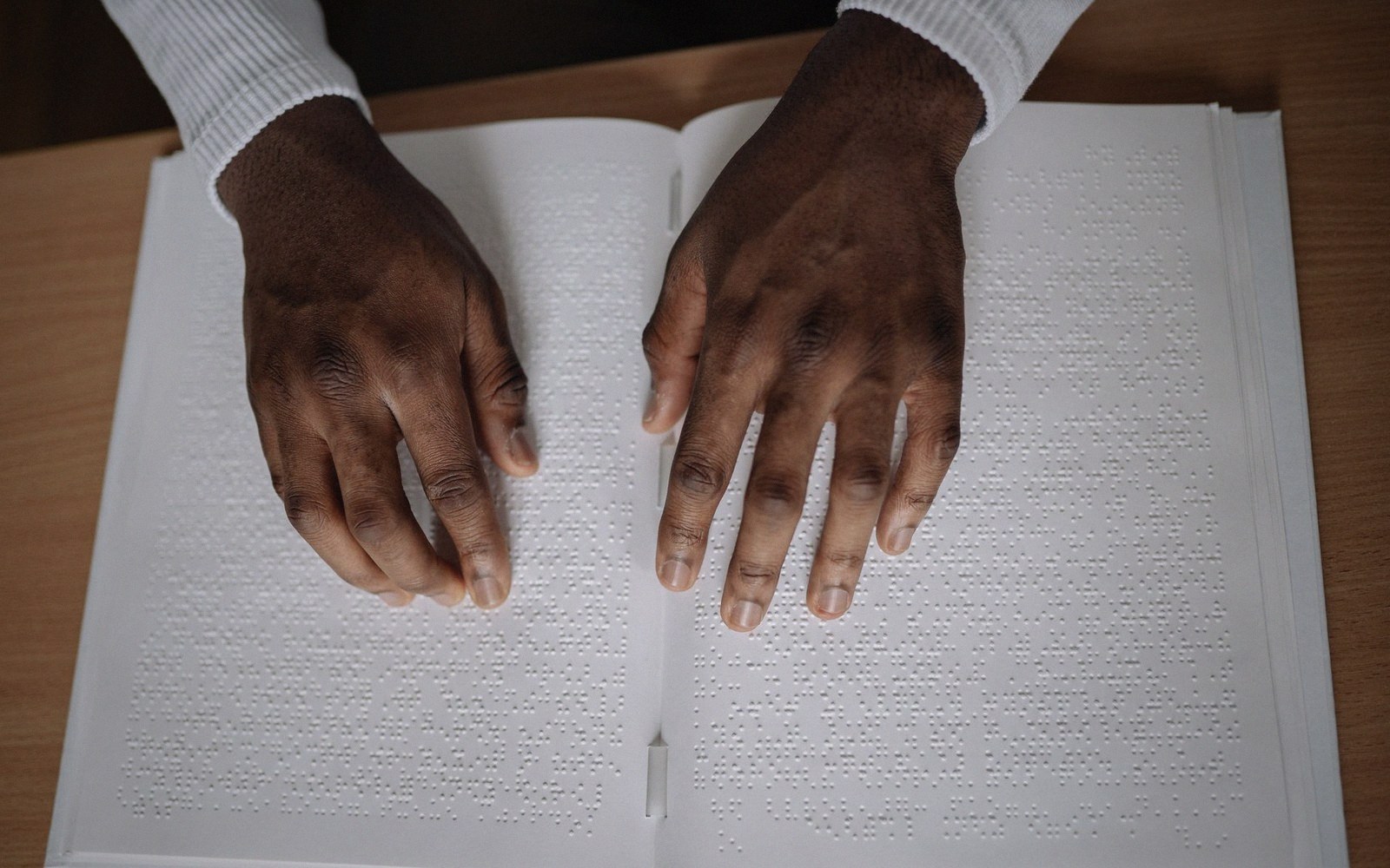 Two hands rest on an open book, fingertips reading raised braille dots on the page