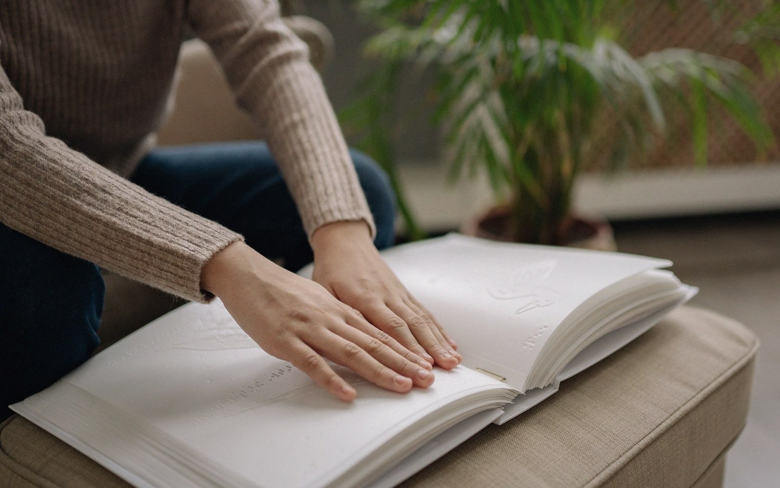 A person reads a braille book with both hands on an ottoman