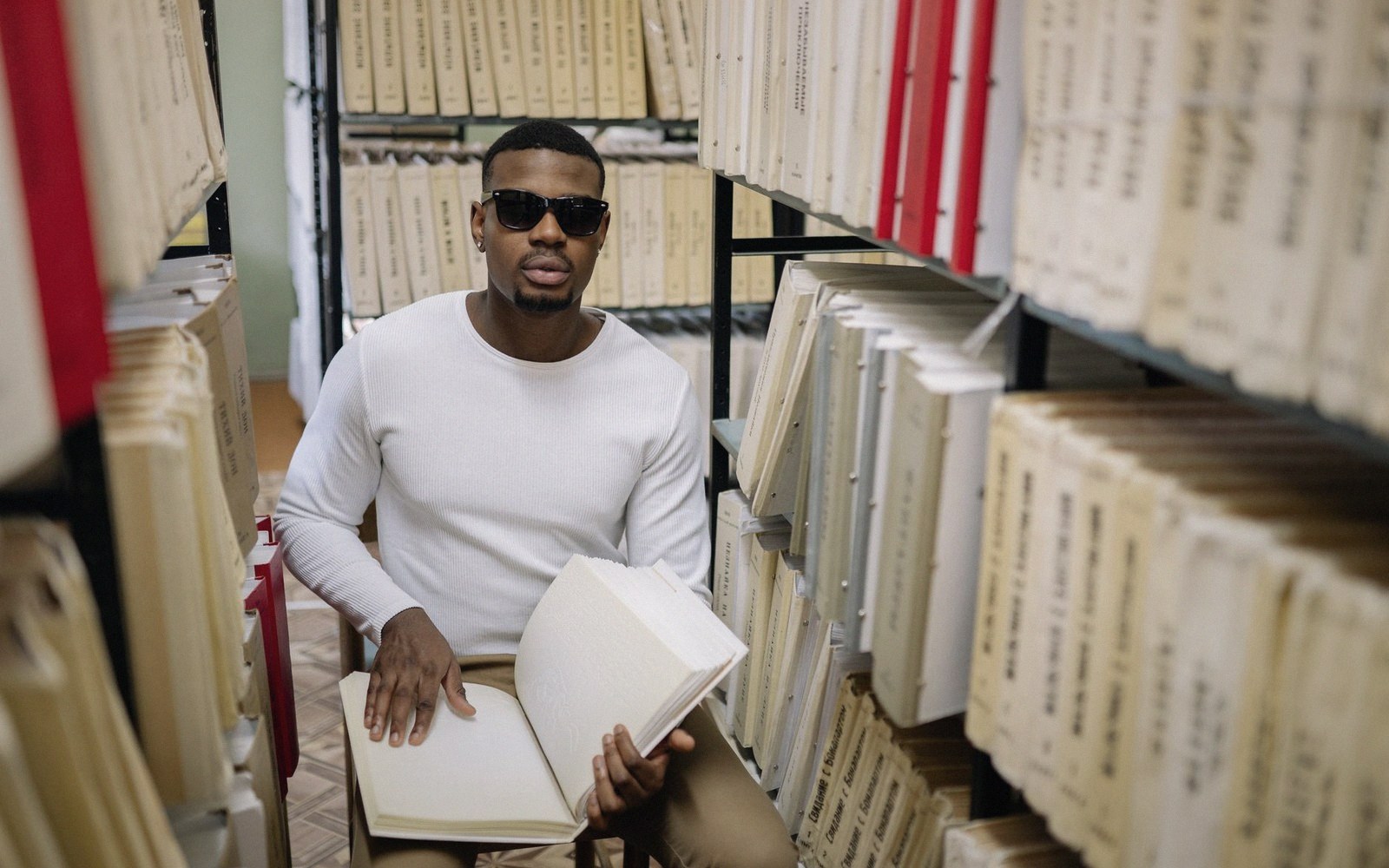 Man in sunglasses sits reading a large bound volume between shelves of archived files