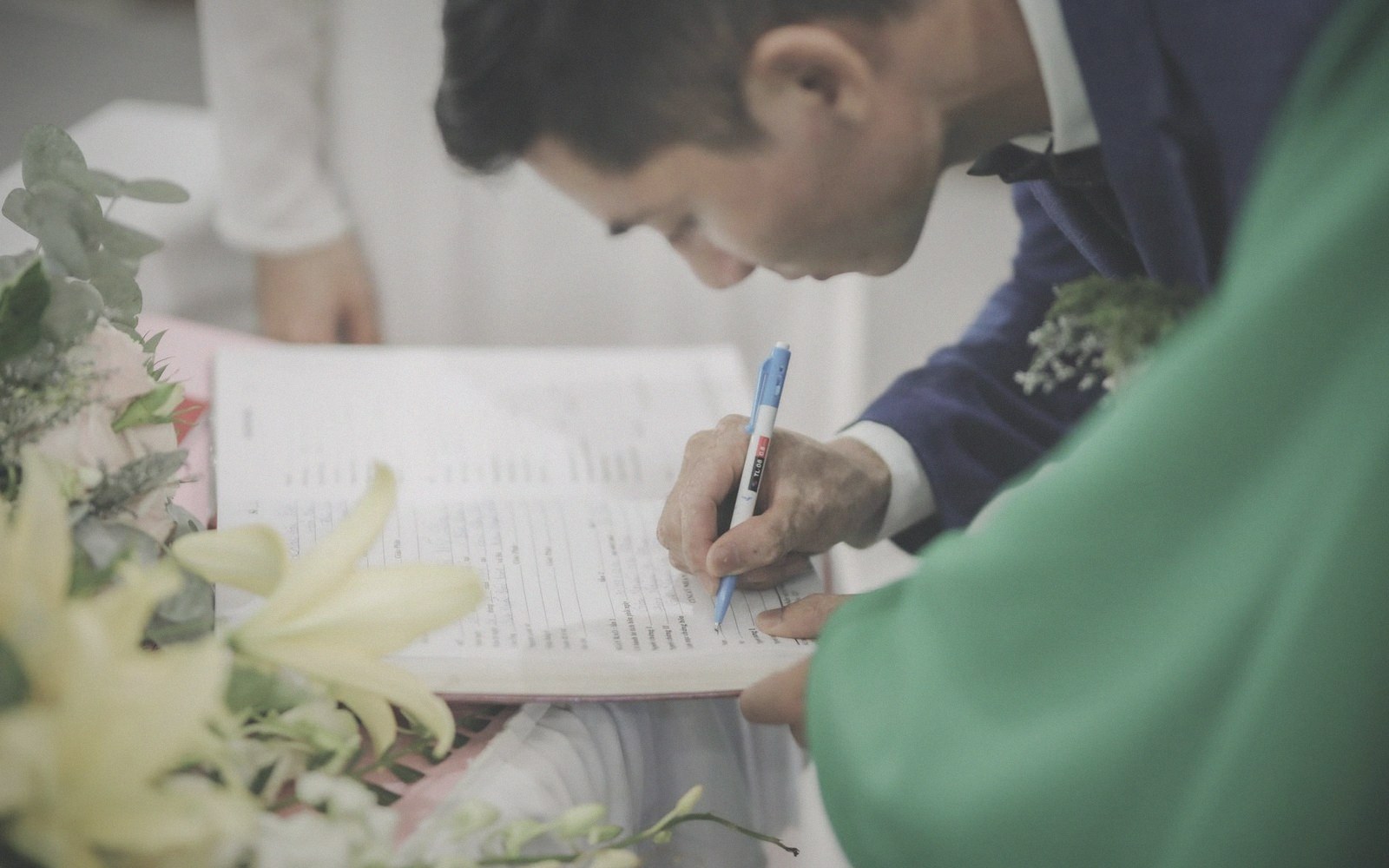 Groom in a navy suit signs a document beside flowers and a priest in green vestments