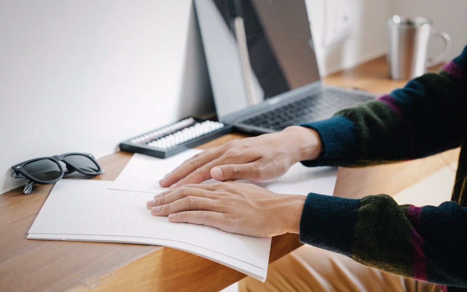 Hands reading a braille document on a desk beside a laptop and abacus