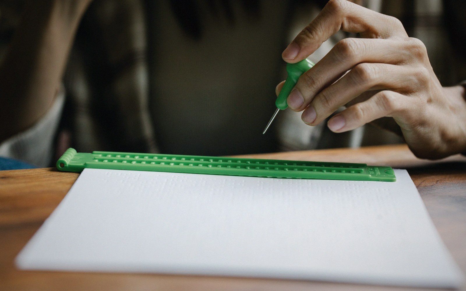 A braille slate and stylus on a desk beside a sheet of heavy paper